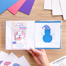 Load image into Gallery viewer, A hand holds an craft package labeled 'Frosty Snowman' 'Card Aperture Pack' 'Embellished edition' on a light wood tabletop. The package contains blue, pink and purple-hat snowman cutout on white cardstock and an iris folding pattern sheet.
