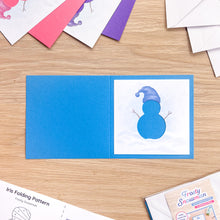 Load image into Gallery viewer, An open blue greeting card on a light wood tabletop, with a printed snowman illustration on the right panel. Partially visible papers and a pattern sheet are arranged around it, suggesting a aperture card pack.
