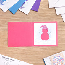 Load image into Gallery viewer, An open pink folding card on a light wood tabletop, showing a snowman illustration on the inside. Partially visible around it are other cards, envelopes, and a folding pattern sheet.
