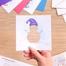 Load image into Gallery viewer, A hand holds a square white card featuring a snowman silhouette with a purple hat over a wood-grain cutout, set on a light wood tabletop with colorful pattern sheets and envelopes around it.
