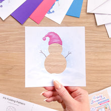 Load image into Gallery viewer, A hand holds a square white paper craft template featuring a snowman silhouette with a pink Santa hat, over a wooden tabletop. Partially visible papers and a printed folding pattern are arranged around it.
