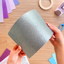 Load image into Gallery viewer, A sheet of light blue glitter card bending so the light reflects off it

