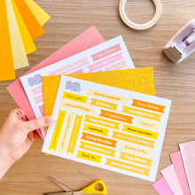 Load image into Gallery viewer, Sheet of printed greetings held by a hand on a wooden surface with stationery items.
