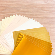 Load image into Gallery viewer, A fanned-out selection of A5 paper sheets in warm tones, including cream, beige, white, sparkle, and metallic gold, arranged on a light wood grain background.

