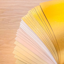 Load image into Gallery viewer, A fanned-out selection of metallic gold, yellow, beige, and white sheets on a light wood grain background.
