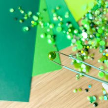 Load image into Gallery viewer, Close-up of green gems and a tweezer tool on a wooden surface with green paper in the background
