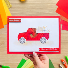 Load image into Gallery viewer, A hand holds a square greeting card with a red border and a red iris fold pickup truck made from layered red paper strips, with small animals in the bed. The card reads 'Happy Birthday' and 'Have a wonderful day' and is photographed on a wooden surface with colorful paper scraps around it.

