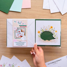 Load image into Gallery viewer, A hand holds a packaged hedgehog card aperture set on a light wood tabletop. The set includes a printed aperture cards, coloured card blanks, envelopes and an iris folding pattern and guide.
