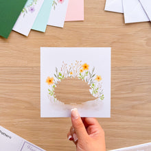 Load image into Gallery viewer, A hand holds a square white card with a cutout design of a hedgehog surrounded by yellow flowers and green leaves, set on a light wood tabletop with craft paper sheets in the background.
