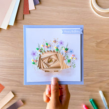Load image into Gallery viewer, A hand holds a square handmade birthday card with a light blue border and an iris fold hedgehog in the center. The card has a small blue banner reading 'Happy Birthday' and is photographed on a light wood desk with craft supplies around it.
