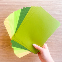 Load image into Gallery viewer, Hand holding a stack of green paper sheets on a wooden surface
