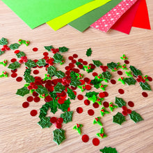 Load image into Gallery viewer, Holly and holly berry craft confetti scattered on a light wood tabletop, with colorful paper sheets visible in the background.
