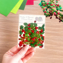 Load image into Gallery viewer, A hand holds a clear plastic packet of glittery 'Holly &amp; Berries Mixed Pack' embellishments, showing green holly leaves and red berries, against a wooden tabletop background with loose pieces scattered.

