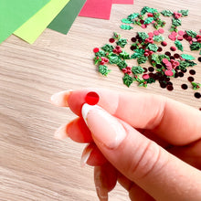 Load image into Gallery viewer, A close-up of a hand with manicured nails holding a small, round, glossy red berry embellishment. In the background, scattered on a light wood surface, are many similar red, green, and dark brown holiday-themed sequins or craft decorations, with colorful paper sheets visible in the upper left corner.
