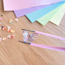 Load image into Gallery viewer, Butterfly-shaped object on a wooden surface with colourful paper in the background.
