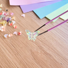 Load image into Gallery viewer, Butterfly-shaped gemstone on tweezers with colourful gemstones and paper in the background.
