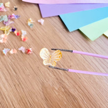 Load image into Gallery viewer, Butterfly-shaped object on a wooden surface with pink tweezers.
