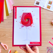 Load image into Gallery viewer, Hand holding an iris fold wine glass greeting card with 'Cheers to you' and 'congratulations' on a wooden surface.
