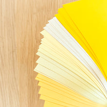 Load image into Gallery viewer, Thirty sheets of A5 paper in 5 different yellow/gold/cream shades fanned out against a wooden surface

