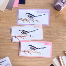 Load image into Gallery viewer, Three handmade bird greeting cards arranged on a wooden table, each featuring an iris fold bird on a branch with pastel borders in purple, blush pink and bright pink. Card decorations and craft supplies are visible around the edges.
