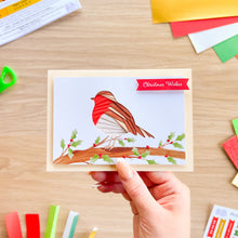 Load image into Gallery viewer, A hand holds a handmade Christmas robin greeting card with a red-breasted bird perched on a branch with holly leaves, set against a light wood desk with colorful craft paper pieces scattered around.
