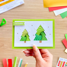 Load image into Gallery viewer, A handmade greeting card held by a hand, featuring two green layered paper Christmas trees on a white background with a bright green border. The card is photographed on a light wood desk with craft supplies scattered around.
