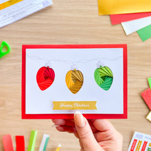 Load image into Gallery viewer, A handmade Christmas greeting card held by a hand, featuring three colorful Christmas lights on a printed silver chain in red, gold, and green on a white background with a red border. The card has a 'Happy Christmas' banner at the bottom and is photographed on a wooden desk with craft supplies scattered around.
