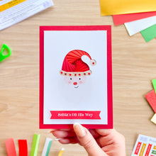 Load image into Gallery viewer, A handmade Christmas greeting card held by a hand, featuring a red border, a white center with a Santa face wearing a red  iris fold hat, and a red banner reading 'Santa's On His Way'. The card is photographed on a light wood desk with craft supplies scattered around.
