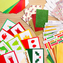 Load image into Gallery viewer, A flat-lay display of a Christmas-themed craft kit on a light wood background, showing colorful cardstock sheets, glitter paper, printed greeting cards, and sticker sheets.
