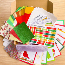 Load image into Gallery viewer, A Christmas card folding kit with various colorful cardstock sheets, envelopes, stickers, and printed greeting cards fanned out inside and around an open cardboard box on a light wood surface.
