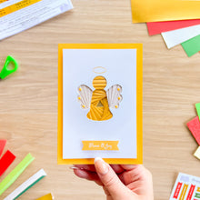 Load image into Gallery viewer, A hand holds a handmade greeting card featuring a geometric yellow and white angel design with a halo and wings, set against a wooden desk with craft supplies scattered around.
