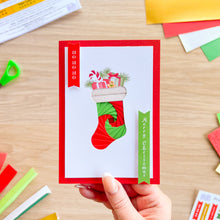Load image into Gallery viewer, A hand holds a Christmas stocking greeting card with a red border and illustration of a red and green stocking filled with gifts. The card is photographed on a light wood desk with colorful paper scraps scattered around.
