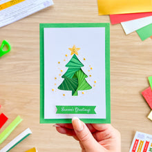 Load image into Gallery viewer, A handmade green Christmas greeting card held by a hand, featuring a layered green Christmas tree with a gold star and the words 'Season's Greetings' on a green banner. The card is set against a light wood desk with colorful craft paper scattered around.
