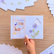 Load image into Gallery viewer, A hand holds a packaged mini butterfly garden aperture card pack on a light wood tabletop. The package contains white butterfly card apertures with printed flower designs.
