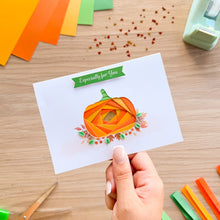 Load image into Gallery viewer, A hand holds a white greeting card featuring an iris fold pumpkin design with the text 'Especially for You' on a green banner. The card is photographed on a wooden desk with craft supplies like colored paper strips, small leaf confetti, and a light green tape dispenser visible around it.

