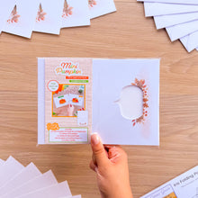 Load image into Gallery viewer, A hand holds a packaged mini pumpkin aperture card pack on a light wood tabletop. The clear plastic package contains printed cards and a small pumpkin-shaped window cutout on the front.
