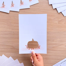Load image into Gallery viewer, A hand holds a white folding card with a die-cut pumpkin design on a light wood tabletop. Similar cards and envelopes are scattered around the surface.

