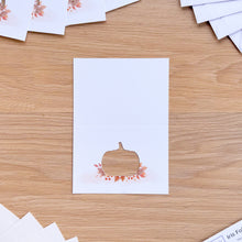 Load image into Gallery viewer, A white rectangular paper card with a pumpkin-shaped cutout and a small autumn floral illustration around the base, placed on a light wood surface with matching papers scattered around the edges.
