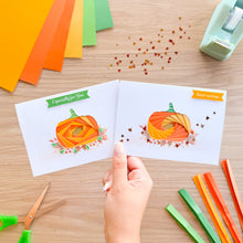 Load image into Gallery viewer, A hand holds two white greeting cards featuring iris fold pumpkin designs in orange, yellow, and green, with autumn leaf sequins. The cards are displayed on a wooden desk surrounded by craft supplies, including colored paper strips, scissors, and a tape dispenser.

