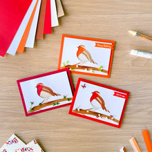 Load image into Gallery viewer, Three handmade greeting cards featuring iris fold robin birds on branches, arranged on a light wood tabletop with craft supplies like colored cardstock, a gem picker tool, and a glue stick nearby.
