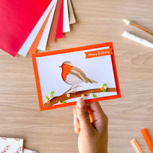 Load image into Gallery viewer, A hand holds a handmade greeting card featuring an iris fold robin bird on a branch, with a 'Happy Birthday' banner. The card is set against a wooden desk with craft supplies like colored paper sheets and markers.
