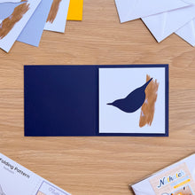 Load image into Gallery viewer, A Nuthatch aperture on a navy blue greeting card blank, displayed on a wooden table with crafting supplies.
