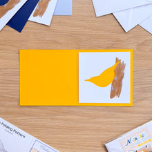 Load image into Gallery viewer, A handmade greeting card shown open on a wooden tabletop. The card has a bright yellow left panel and a white right panel featuring a yellow bird silhouette layered over a brown textured branch shape.

