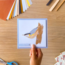 Load image into Gallery viewer, A hand holds a square greeting card featuring an iris fold nuthatch bird, set against a light wood desk with craft supplies in the background.
