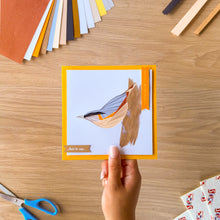 Load image into Gallery viewer, A hand holds a square iris fold greeting card with a bright yellow border and an illustration of a nuthatch perched on a branch. The card is photographed from above on a wooden desk with craft supplies, including colored paper sheets and scissors visible around it.
