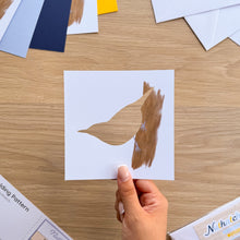 Load image into Gallery viewer, A hand holds a square white card featuring a bird silhouette filled with a brown watercolour tree trunk embellishment, photographed over a wooden tabletop with craft pattern sheets and colored paper visible around it.
