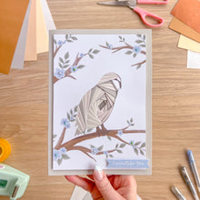 Load image into Gallery viewer, Hand holding a greeting card with a snowy owl design on a wooden table with craft supplies.
