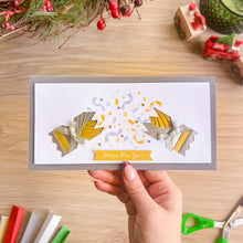 Load image into Gallery viewer, A hand holds a handmade New Year's  card featuring two cracker halves with gold and silver confetti, and a 'Happy New Year' banner. The card is set against a wooden tabletop decorated with festive ornaments.
