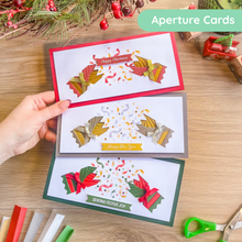 Load image into Gallery viewer, Three handmade iris fold greeting cards are displayed on a wooden table, with a hand holding them. The cards feature layered paper cutouts of pulled crackers, festive ribbons, and confetti, with text banners reading 'Happy Christmas,' 'Happy New Year,' and 'Sending Festive Joy.
