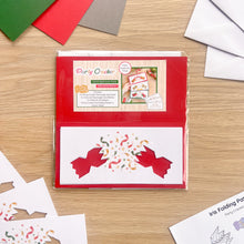Load image into Gallery viewer, Party Cracker card aperture pack in poly bag packaging, displayed on a light wood tabletop with some craft papers and envelopes around it. The pack includes party cracker aperture designs on white cardstock with coloured card blanks and envelopes.
